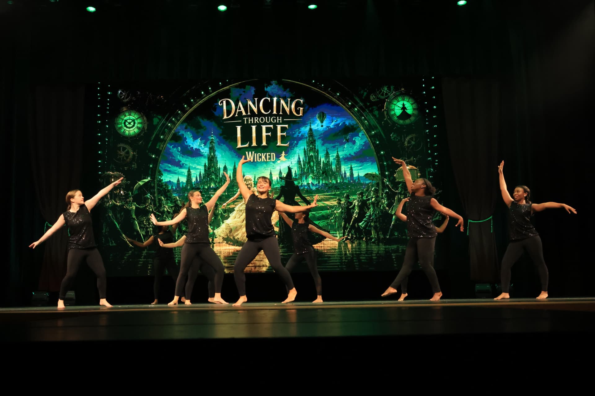 Company dancers in black performing in front of a Dancing Through Life Wicked backdrop