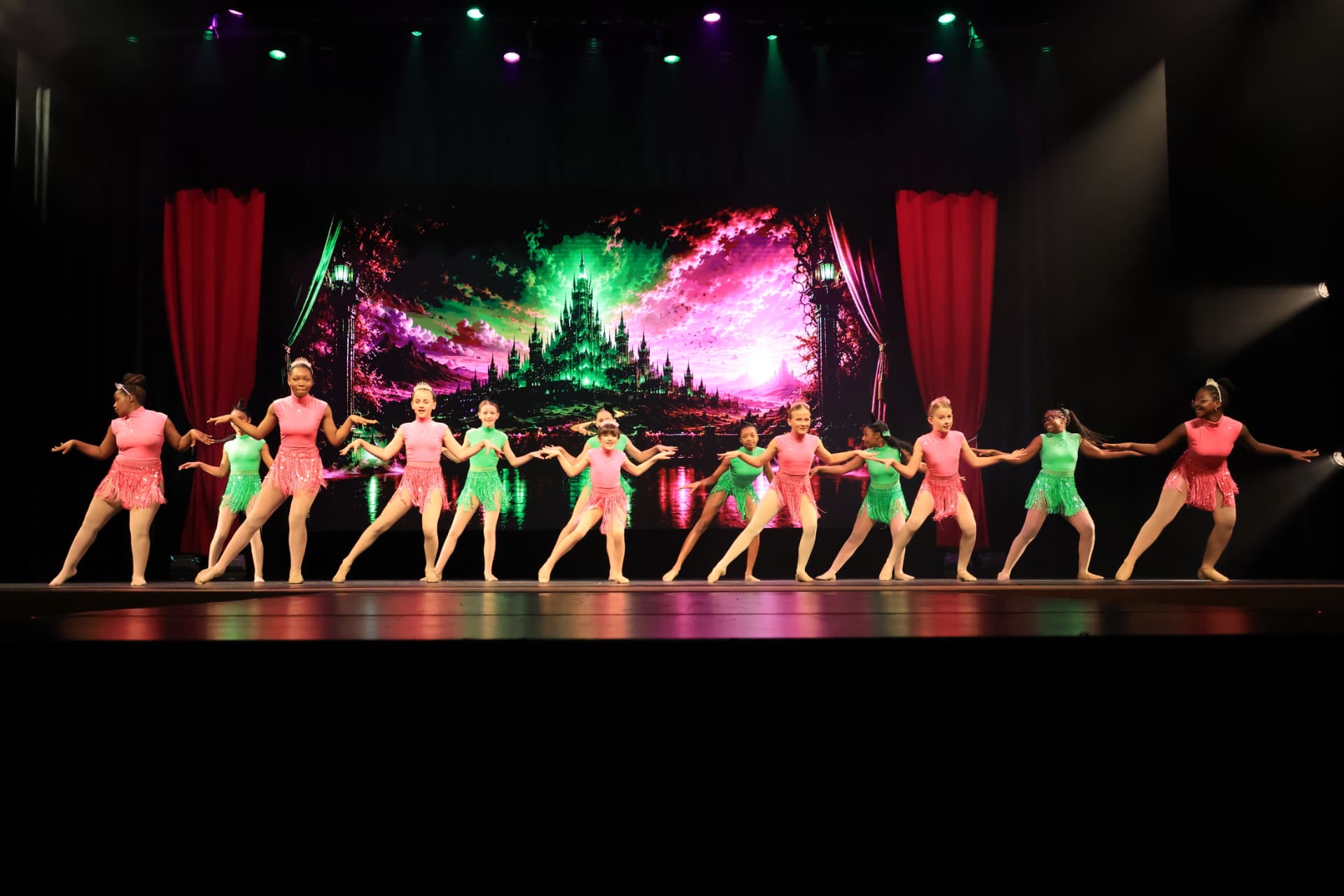 The Company in pink and green fringe costumes in front of an illuminated castle backdrop