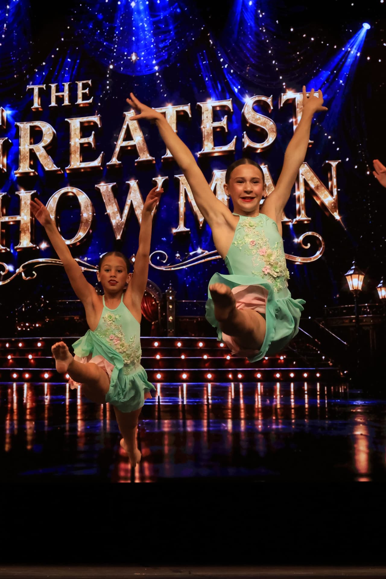 Two dancers leaping in mint green dresses in front of a The Greatest Showman backdrop
